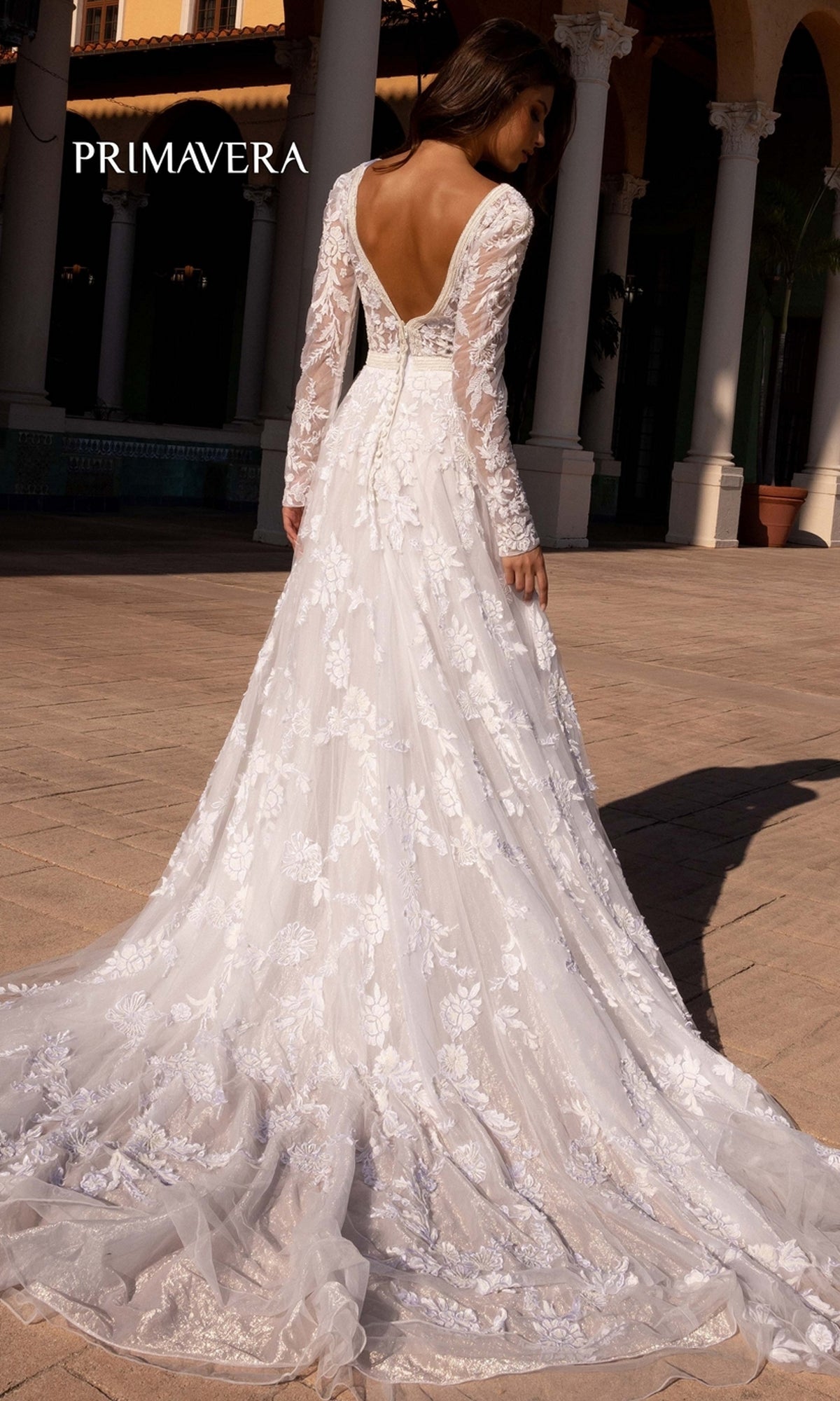 A woman poses outdoors by columns in the Long Wedding Dress 11111 by Primavera, an ivory lace gown with long sleeves and a deep V-back.